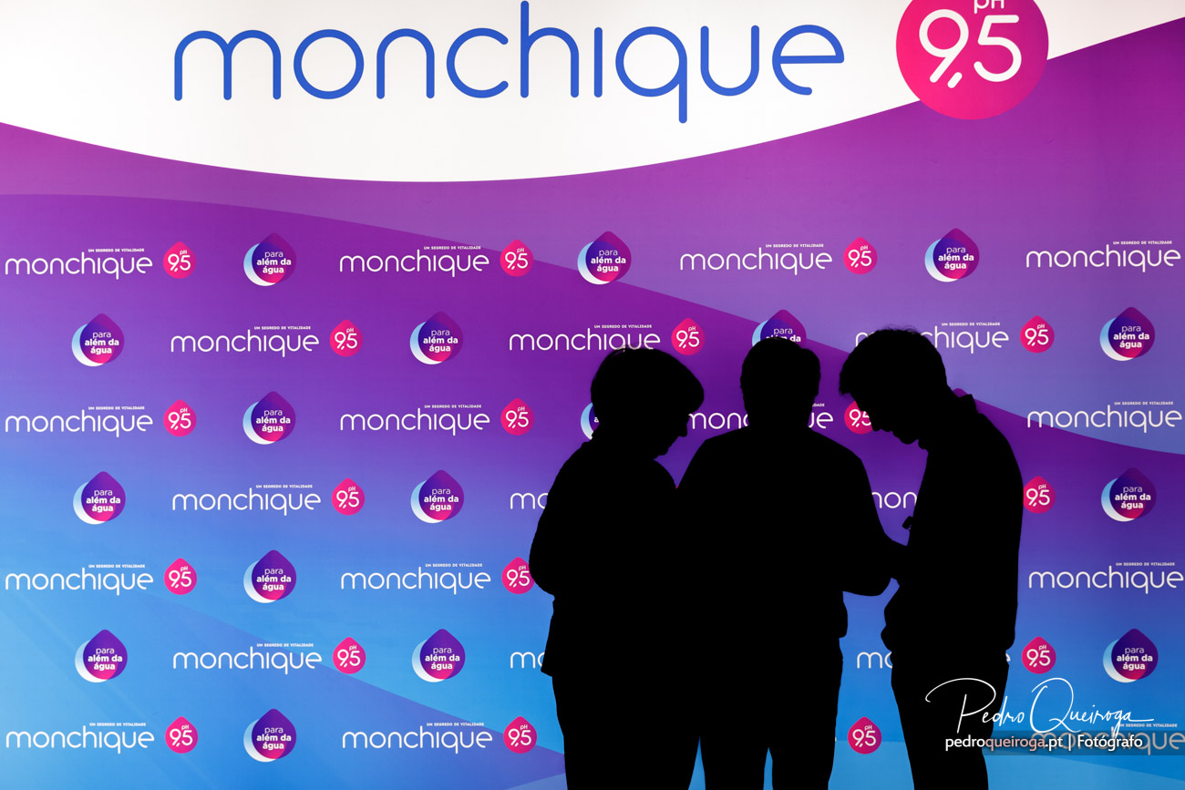 Silhuetas de três pessoas em contraluz diante de painel corporativo com logótipos repetidos em tons roxo e azul