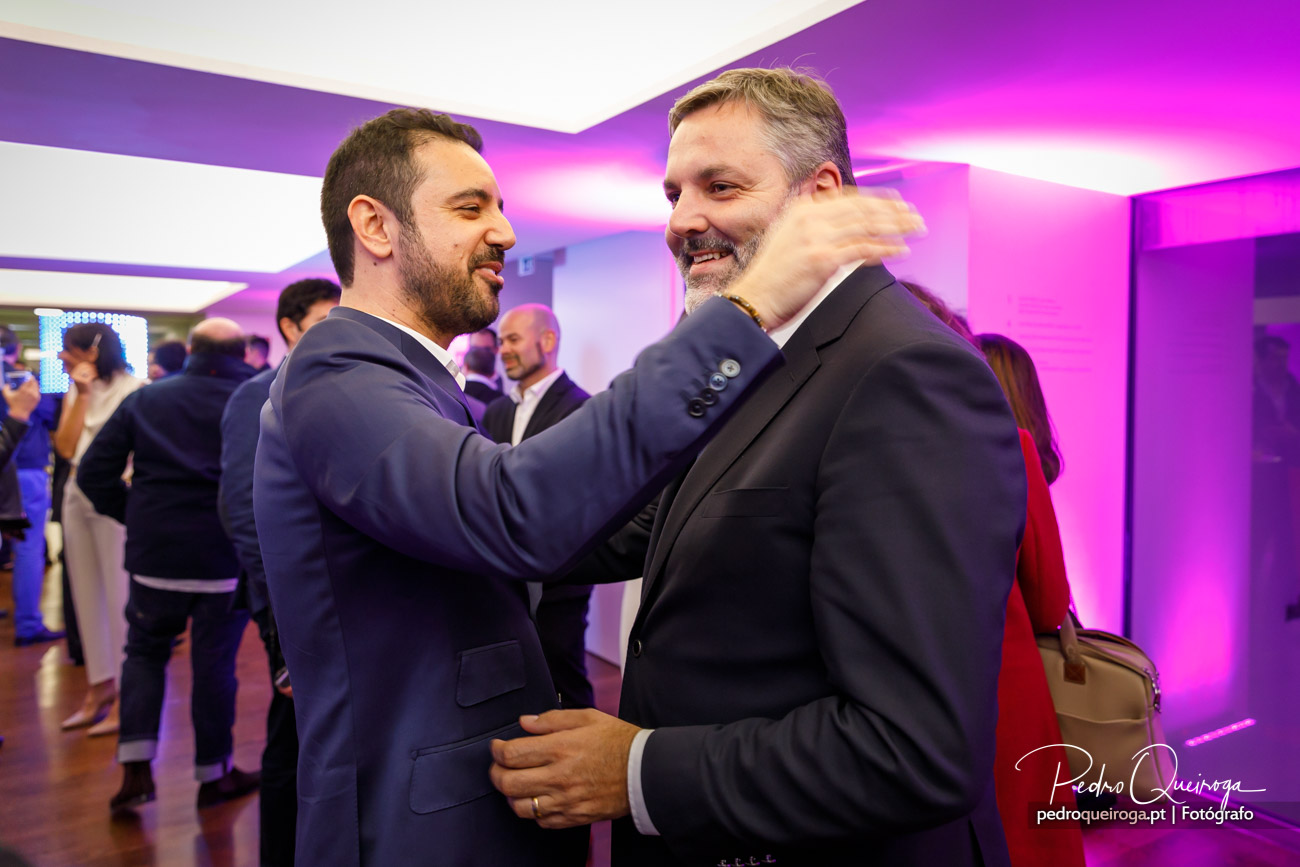 Dois homens de fato cumprimentam-se calorosamente num evento corporativo com iluminação roxa ambiente