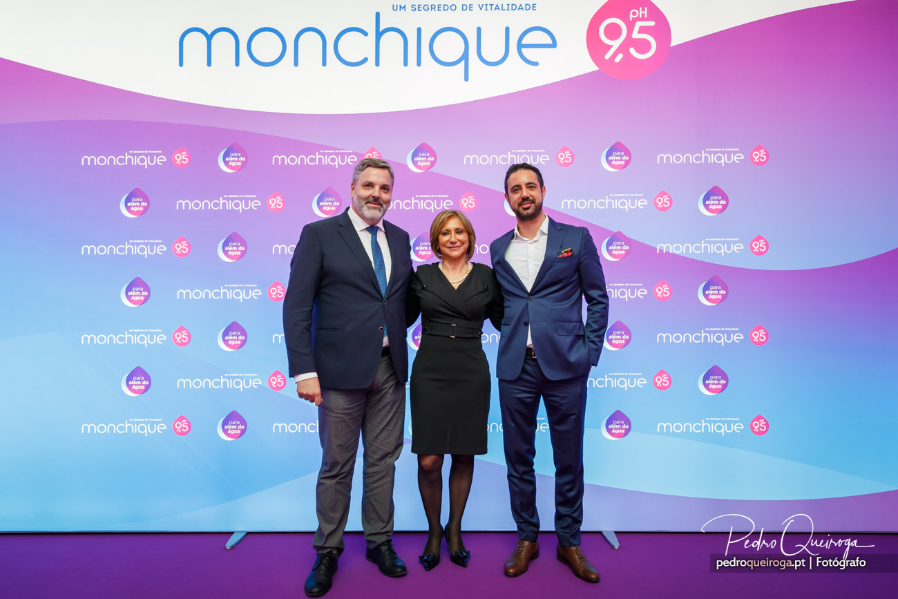 Três executivos posam em photocall da Monchique com fundo degradé azul e rosa num evento corporativo elegante