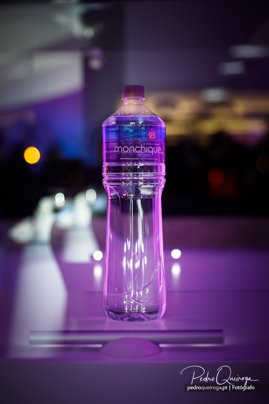 Garrafa de água Monchique iluminada por luz roxa sobre mesa, com fundo desfocado e bokeh dourado