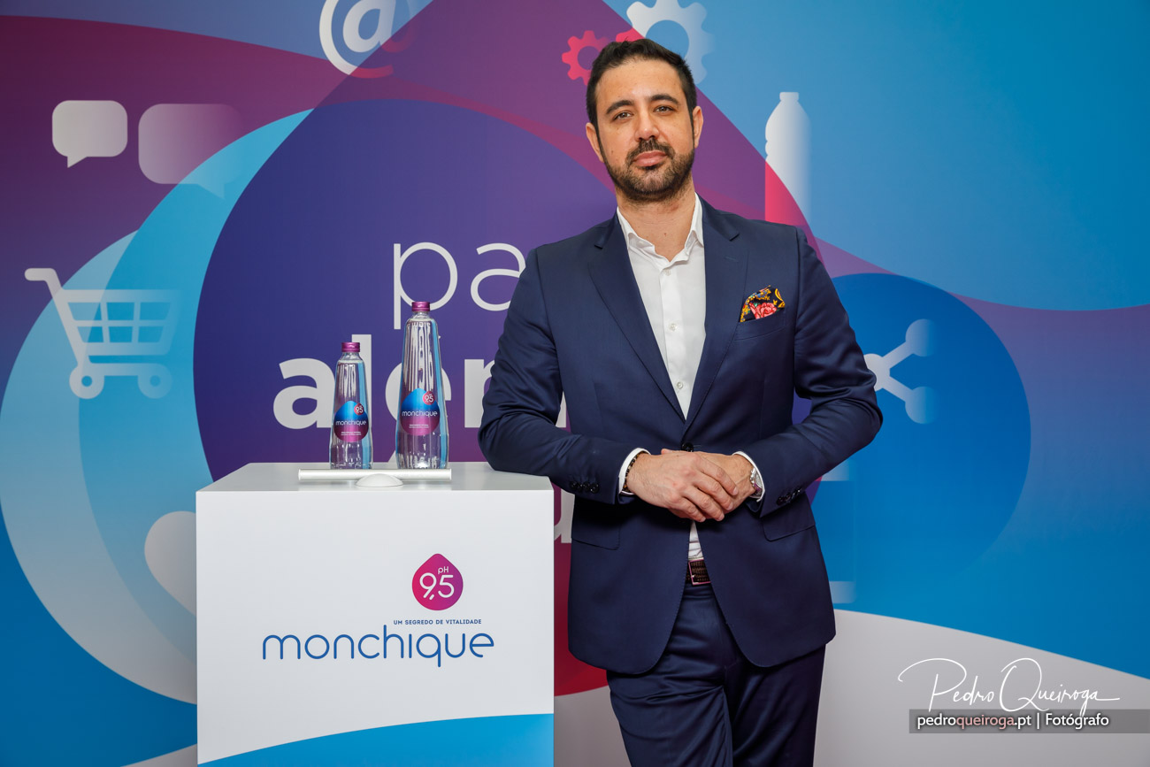 Homem de fato azul junto a púlpito branco com garrafas de água Monchique, sob painel corporativo colorido com ícones digitais.