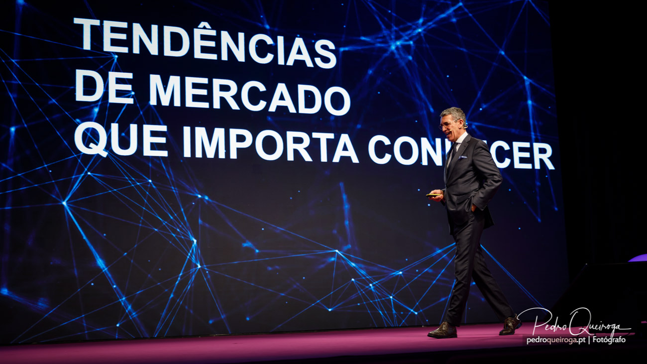 Orador de fato preto caminha sobre palco rosa com ecrã gigante a exibir texto sobre tendências de mercado e fundo tecnológico azul