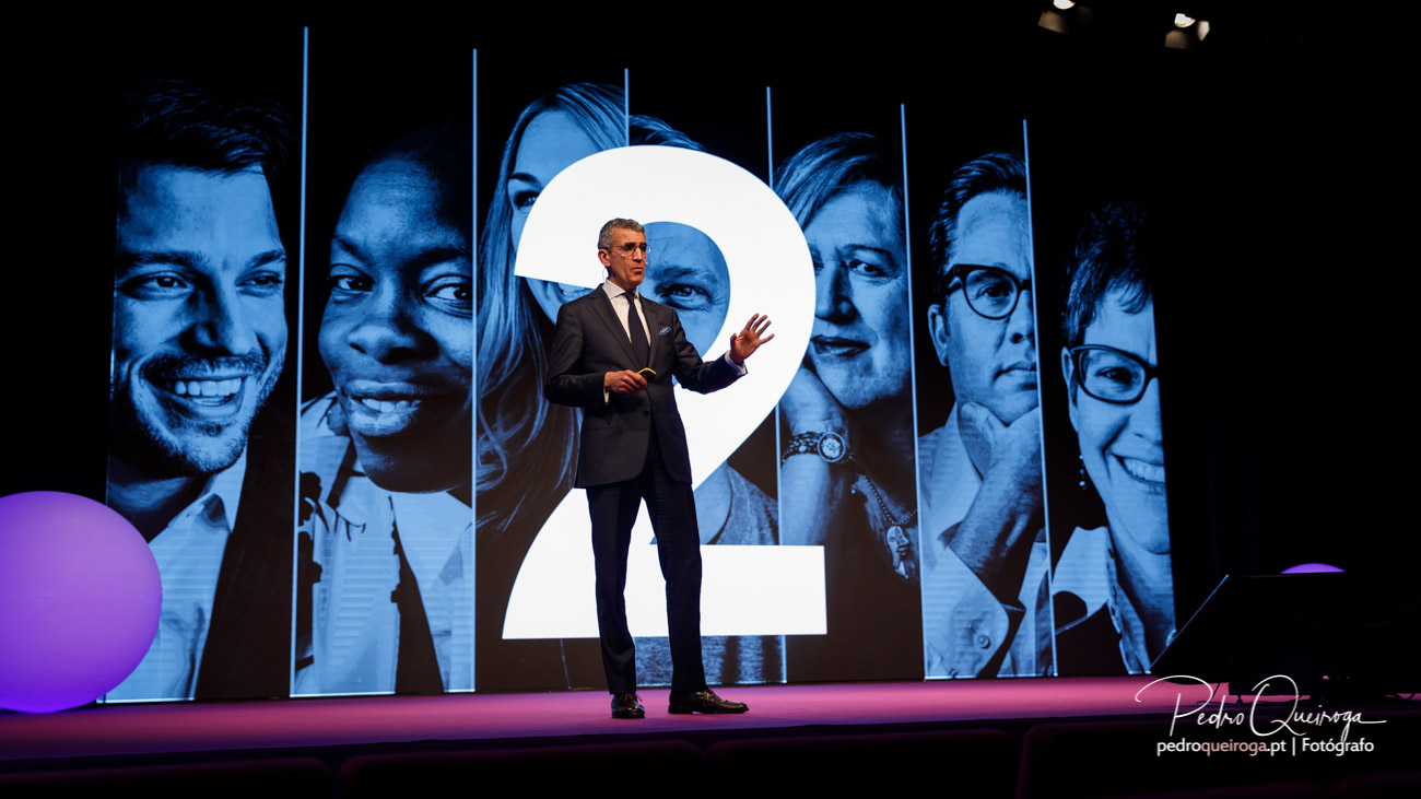 Orador de fato apresenta no palco iluminado com projeção de rostos em tons azuis e número dois ao fundo