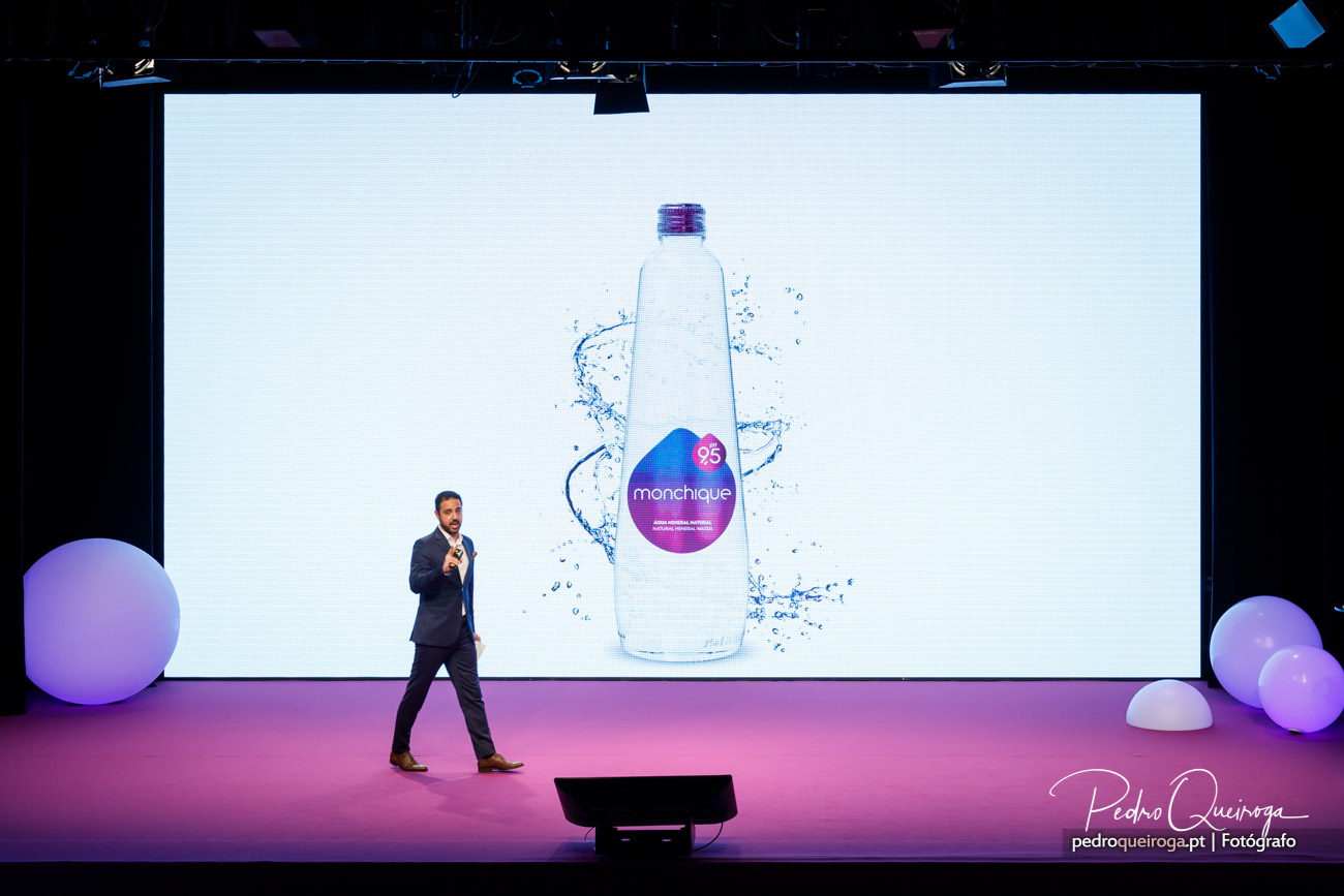 Orador apresenta garrafa de água Monchique num evento corporativo no Algarve com cenário roxo vibrante