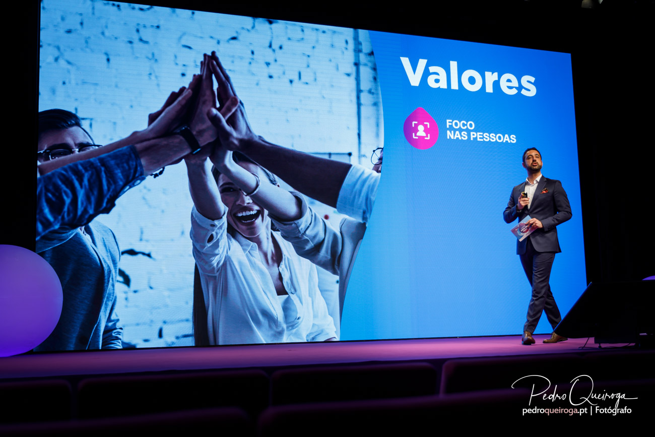 Orador em palco apresenta valores corporativos com fotografias de equipa celebrando união no ecrã gigante