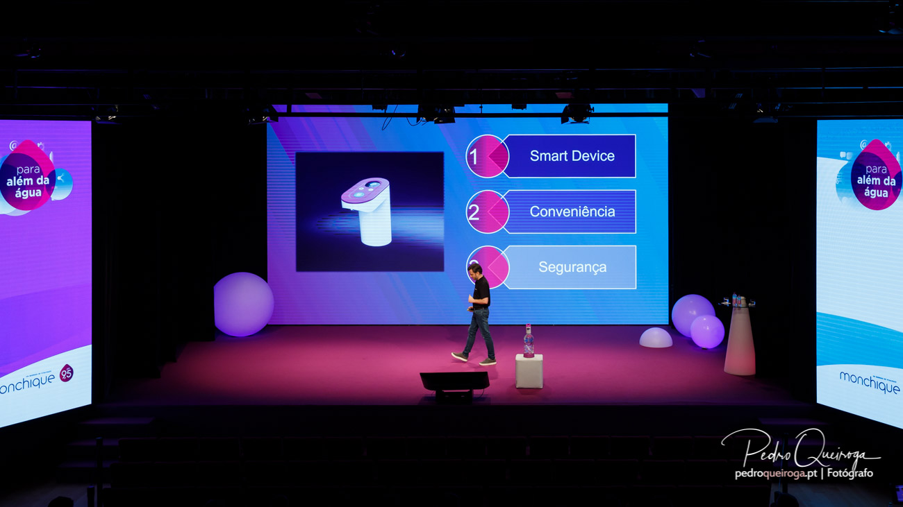 Apresentação corporativa num palco com iluminação rosa e azul, ecrã gigante e elementos decorativos esféricos luminosos