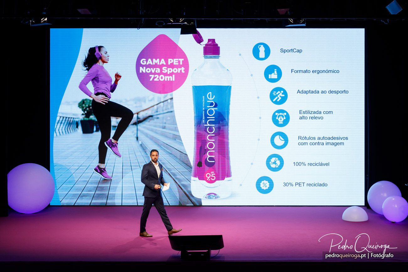 Apresentador em palco corporativo com ecrã gigante a exibir garrafa desportiva Monchique e atleta em movimento
