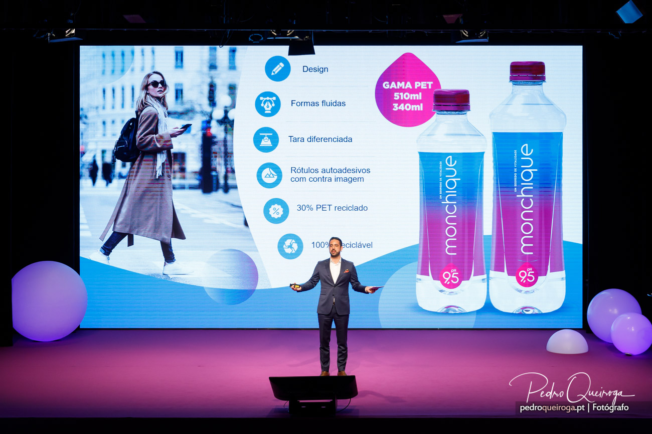 Apresentador em palco corporativo com ecrã a exibir garrafas de água personalizadas e características do produto.