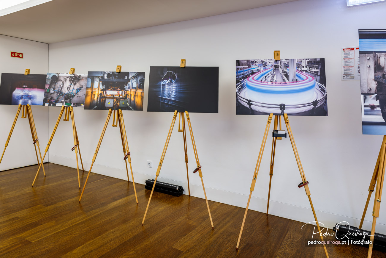 Exposição de fotografia industrial em sala com várias imagens montadas em tripés amarelos sobre pavimento de madeira.