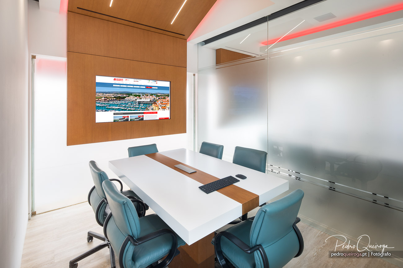 Sala de reuniões moderna com mesa branca, cadeiras azuis, painel de madeira com ecrã e iluminação LED integrada