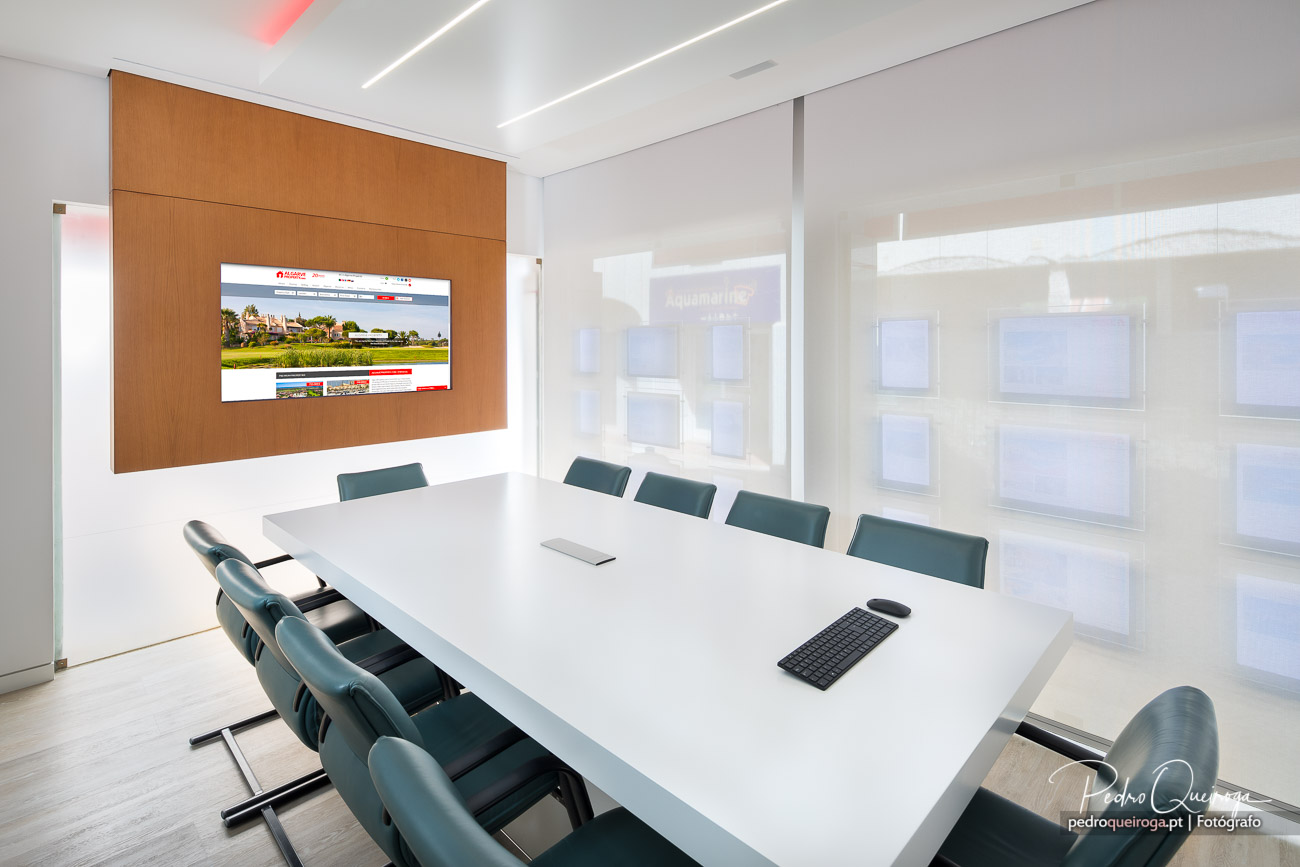 Sala de reuniões moderna com mesa branca, cadeiras verdes e painel de madeira com ecrã no escritório luminoso