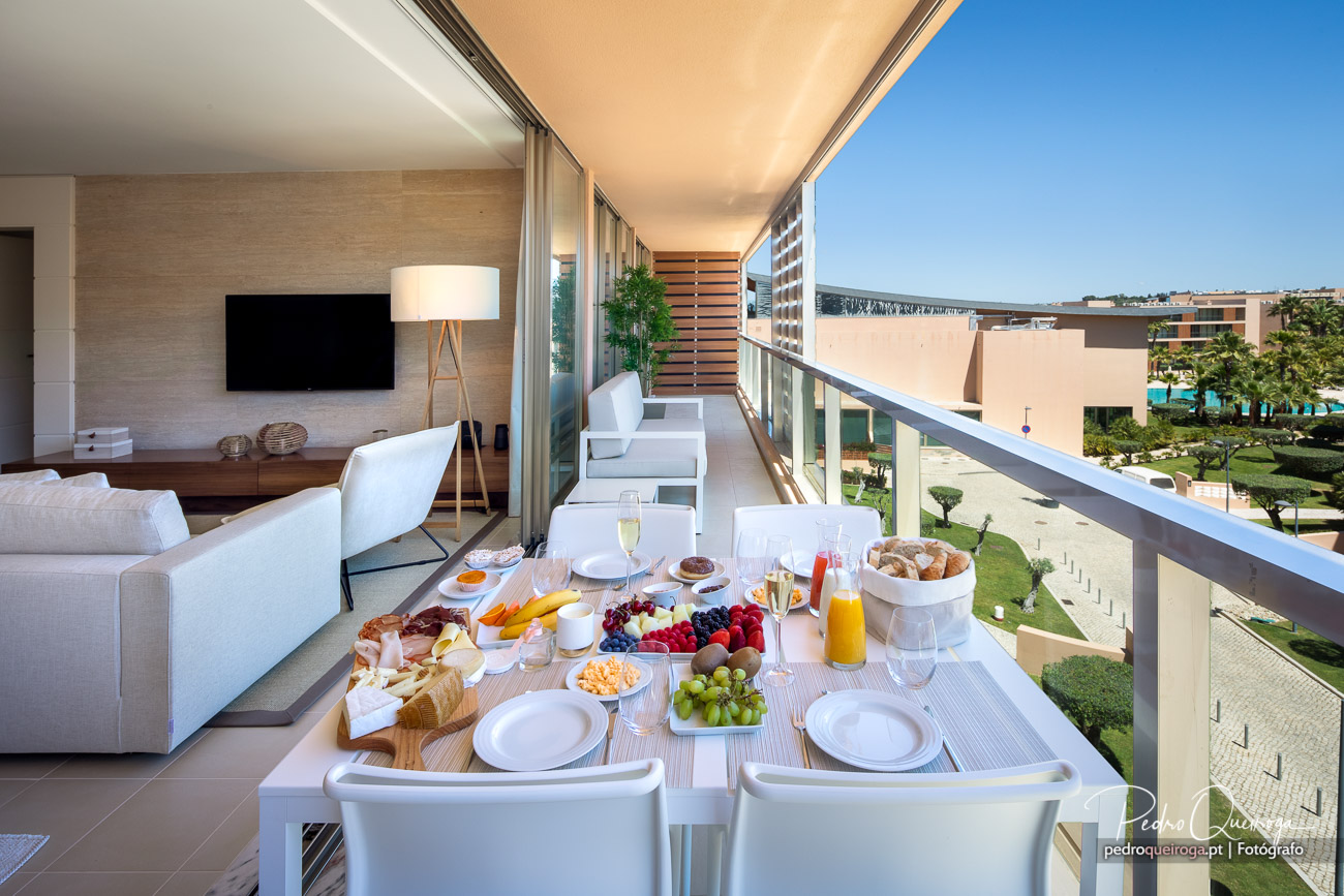 Varanda moderna com pequeno-almoço servido, mobiliário branco e vista para jardim e resort ensolarado ao fundo