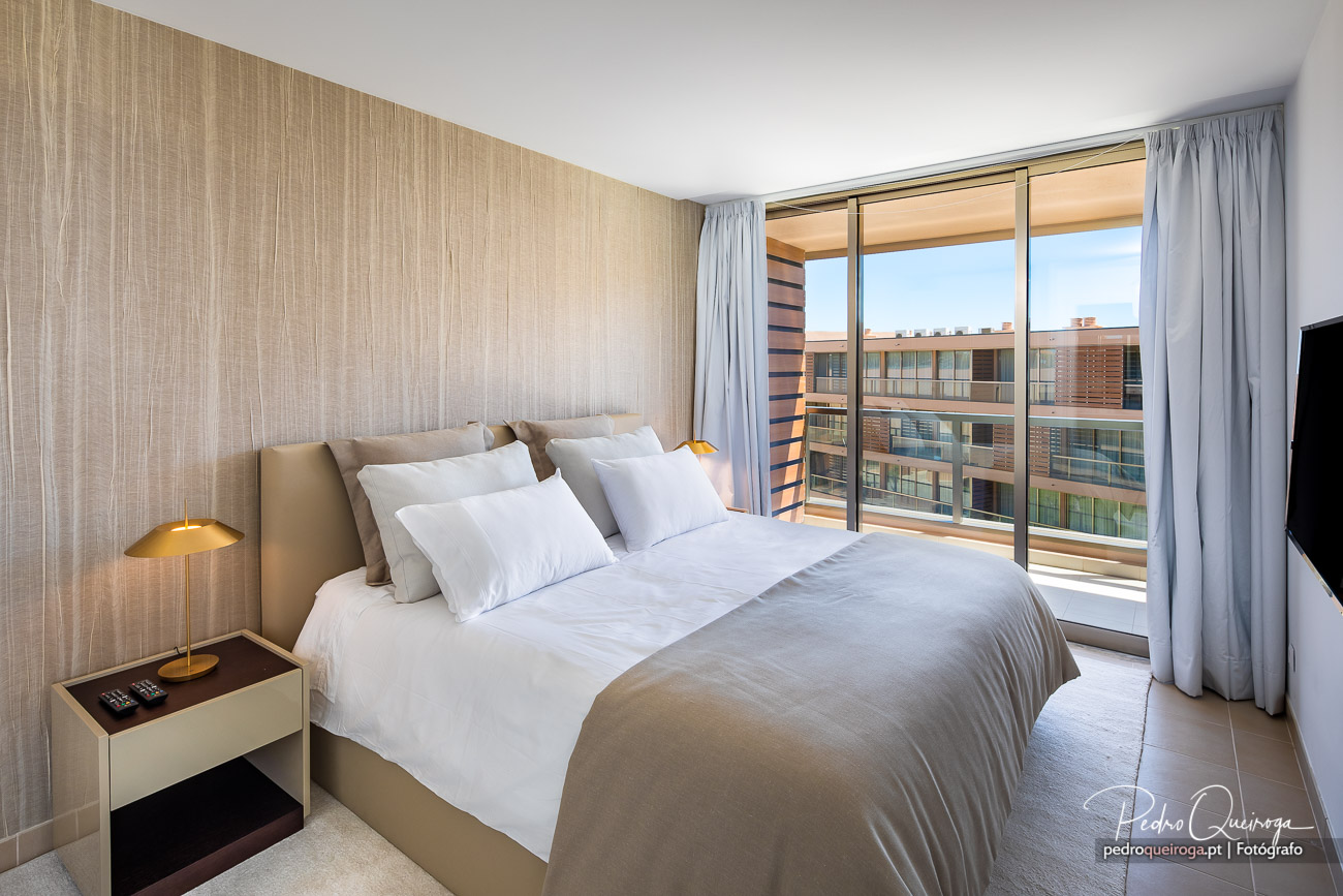 Quarto luminoso com cama de casal, tons neutros e varanda ampla com vista para edifício residencial contemporâneo