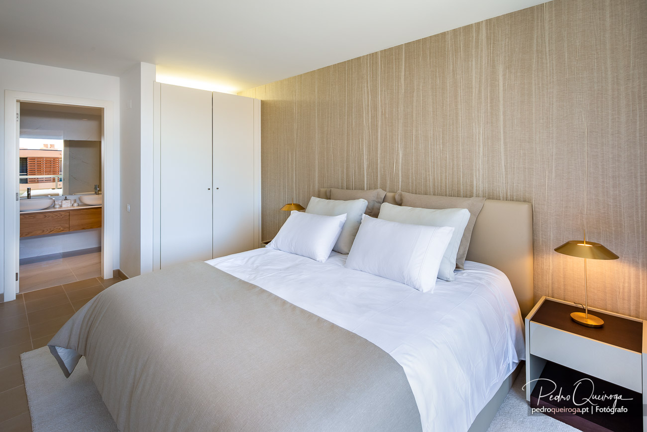 Quarto moderno com tons bege e branco, painel texturizado, casa de banho integrada e iluminação quente e acolhedora.