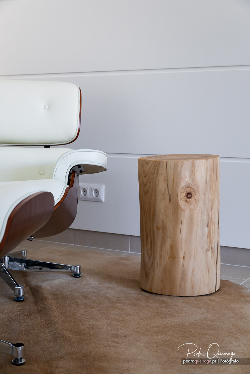 Cadeira branca moderna com detalhes em madeira ao lado de tronco natural sobre pavimento em tons claros e parede neutra