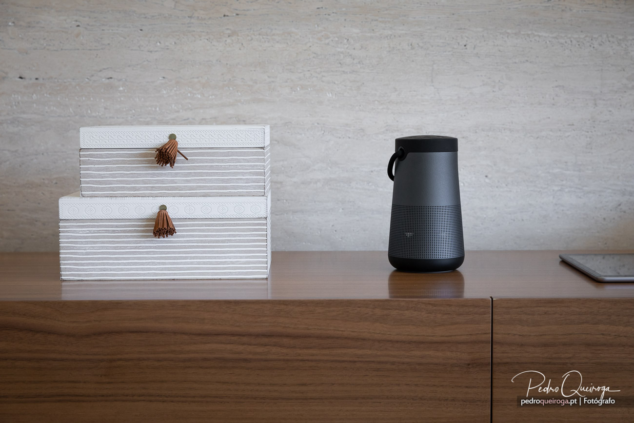 Duas caixas brancas com detalhes em borla castanha e coluna portátil preta sobre mesa de madeira com parede cinza ao fundo