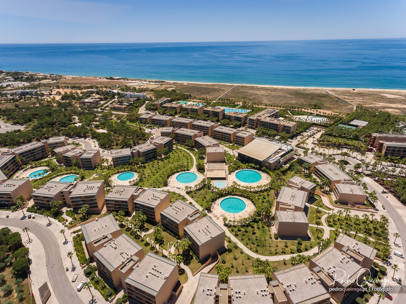 Vista aérea de resort turístico com várias piscinas, edifícios modernos e jardins junto à praia de areia dourada.