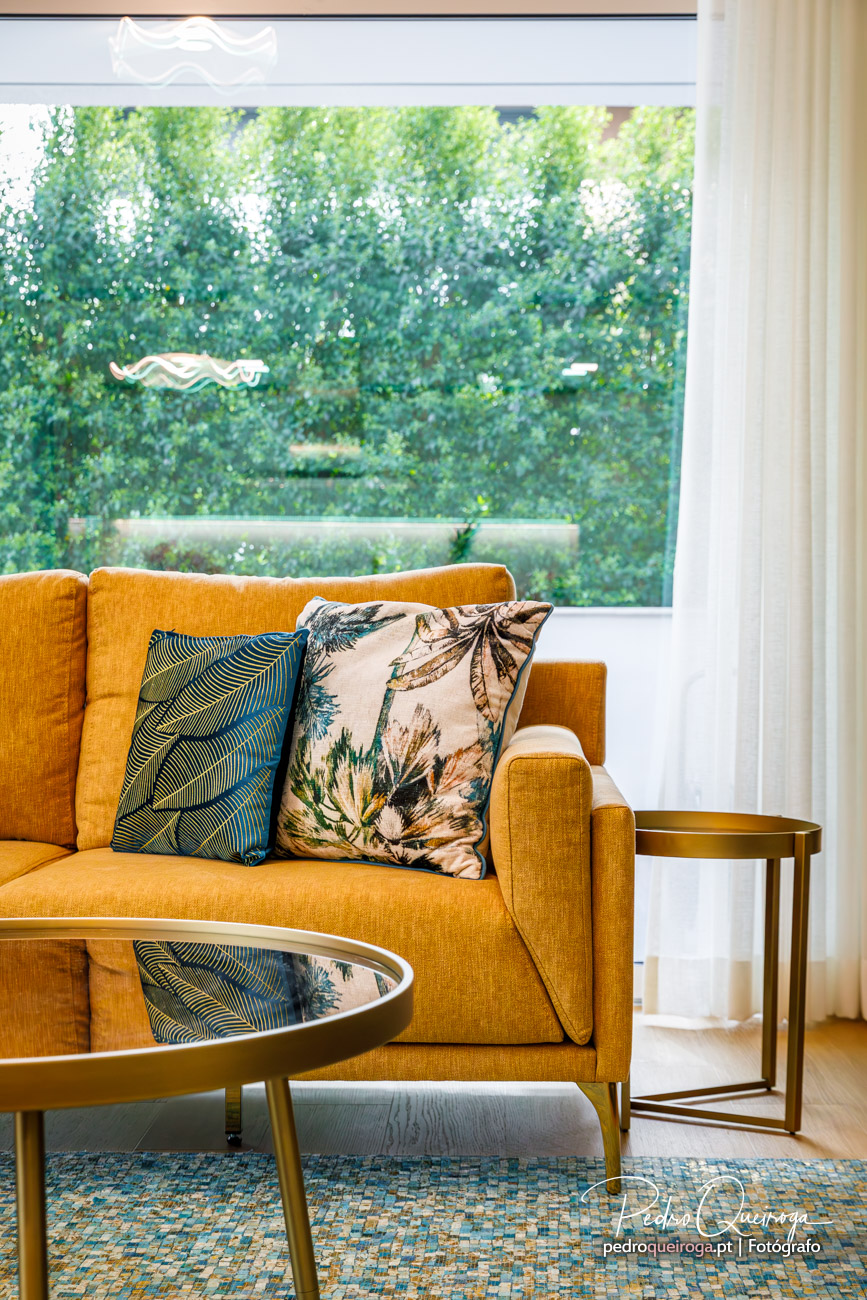 Sofá mostarda com almofadas tropicais junto à janela, numa composição fotográfica de interiores acolhedora e luminosa.