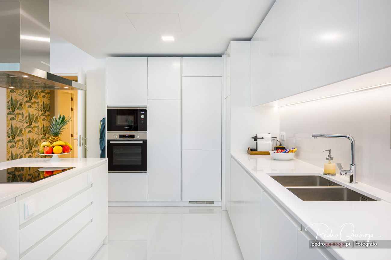 Cozinha moderna branca com eletrodomésticos encastrados, bancada ampla, lava-louça dupla e papel de parede tropical colorido