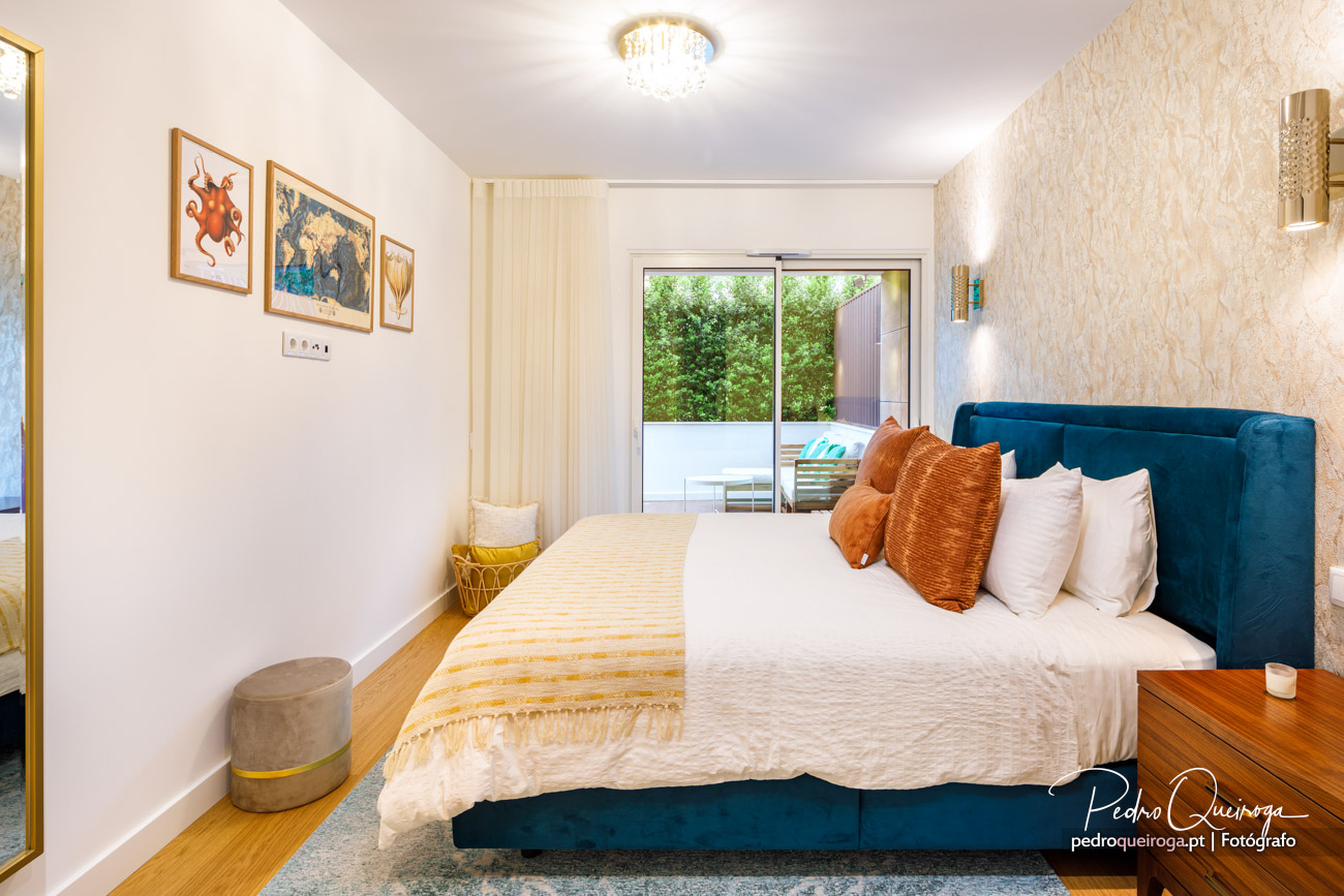 Quarto luminoso com cama azul-petróleo, almofadas laranja e bege, papel de parede texturado e varanda com vegetação.