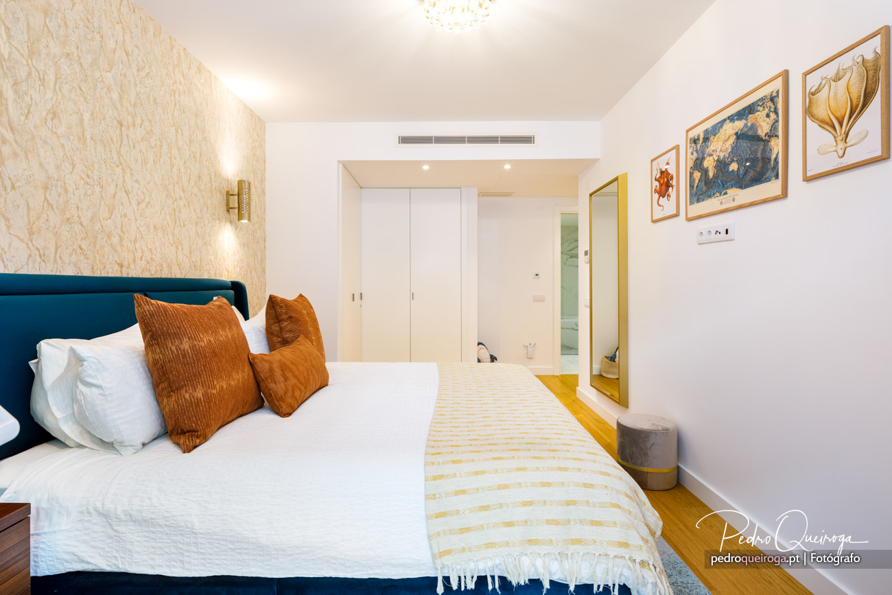 Quarto moderno com cabeceira azul-petróleo, almofadões laranjas, espelho dourado e quadros decorativos na parede branca.