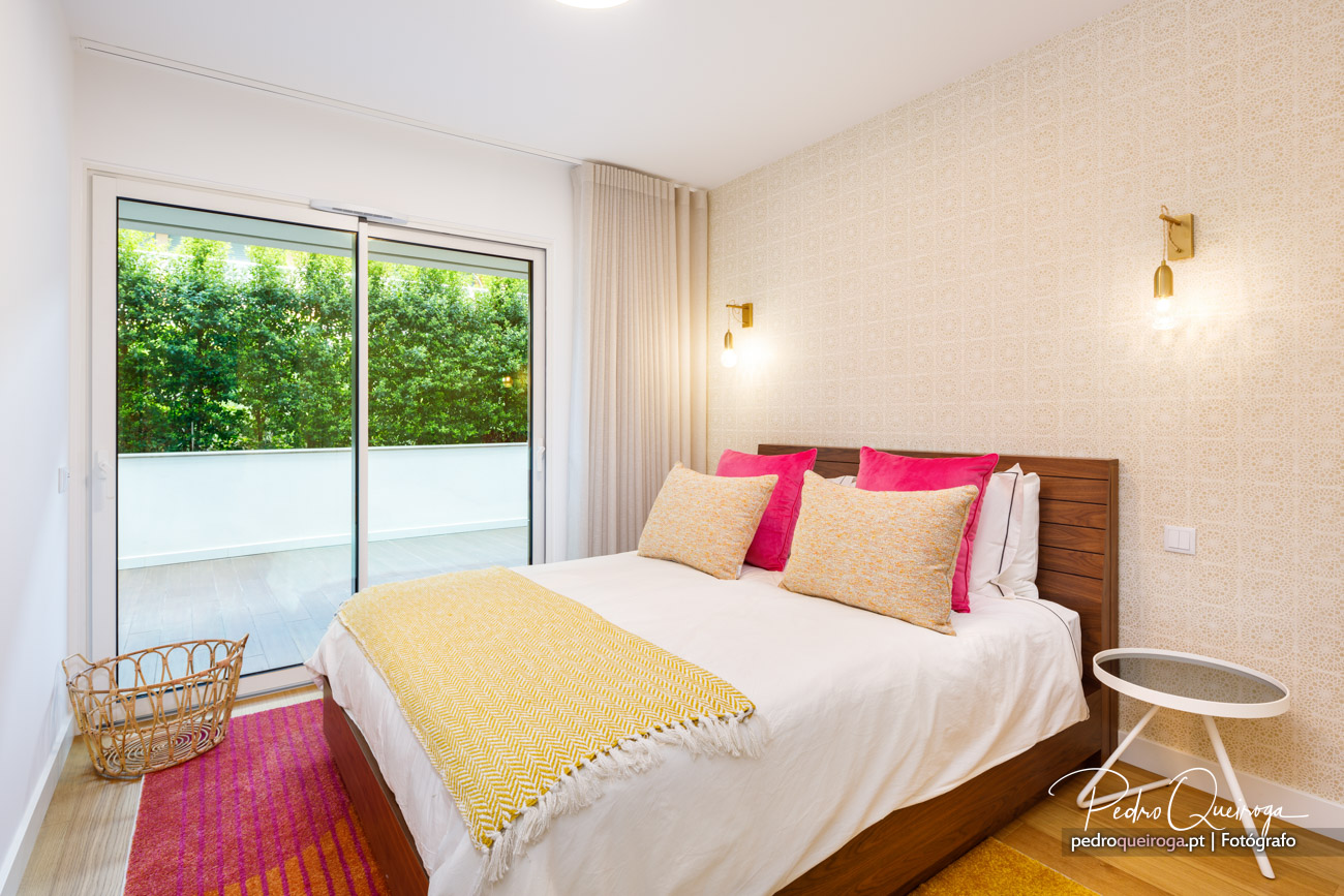 Quarto luminoso com cama de madeira, almofadas rosa e bege, manta amarela e porta de varanda com vista para jardim