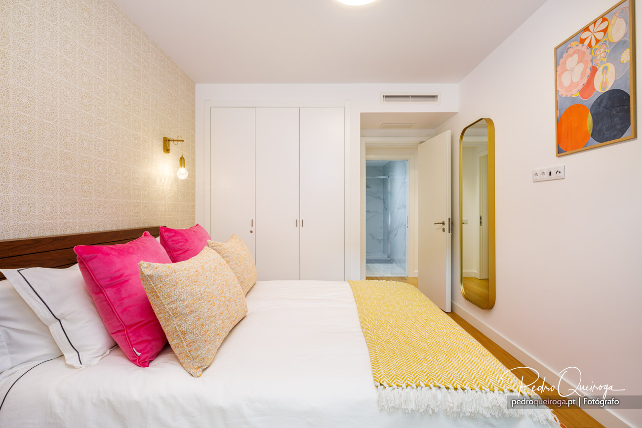 Quarto luminoso com cama de almofadas rosa e amarela, espelho dourado e roupeiro branco embutido