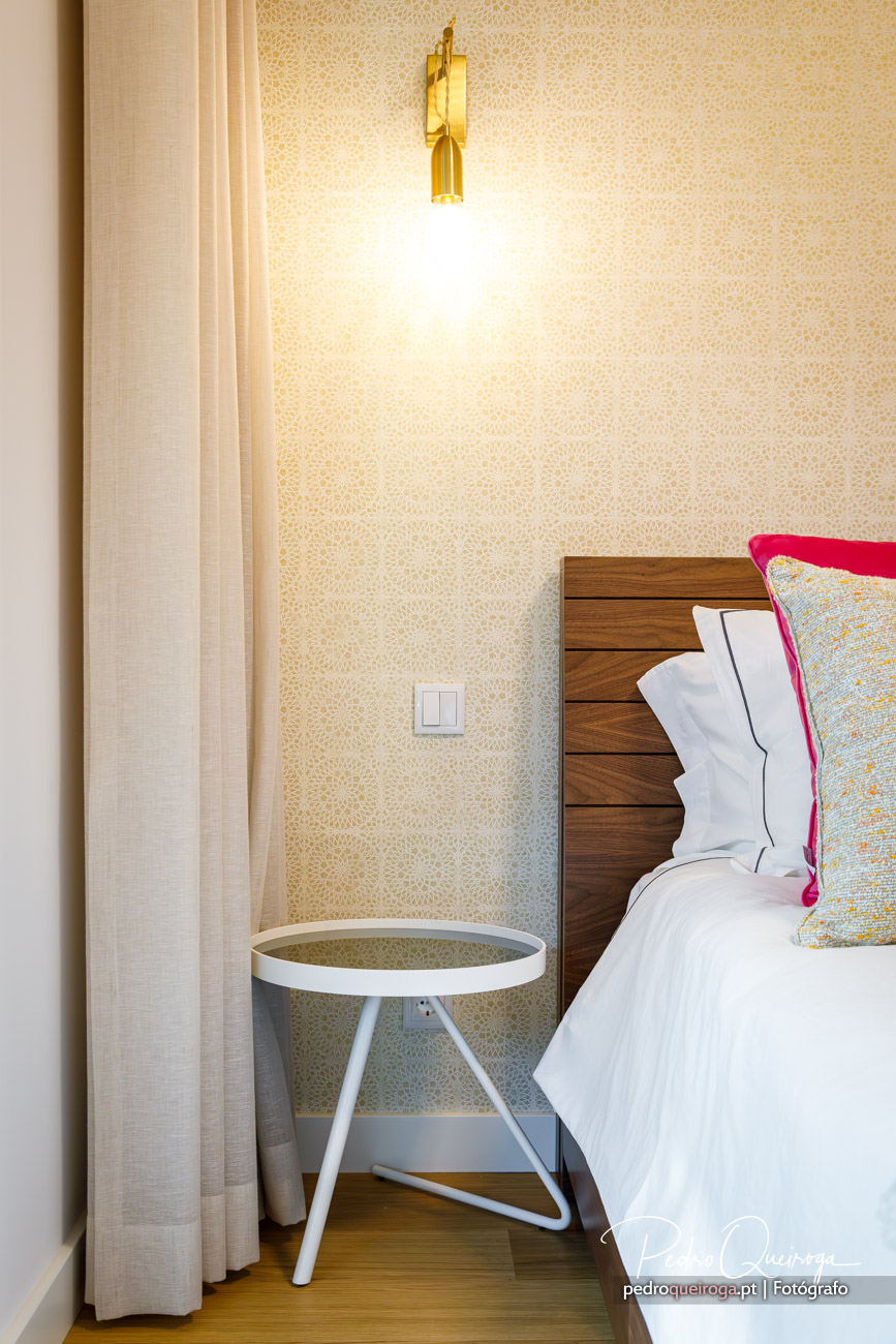 Quarto acolhedor com papel de parede dourado, candeeiro art déco, cabeceira em madeira e almofadas coloridas vibrantes.