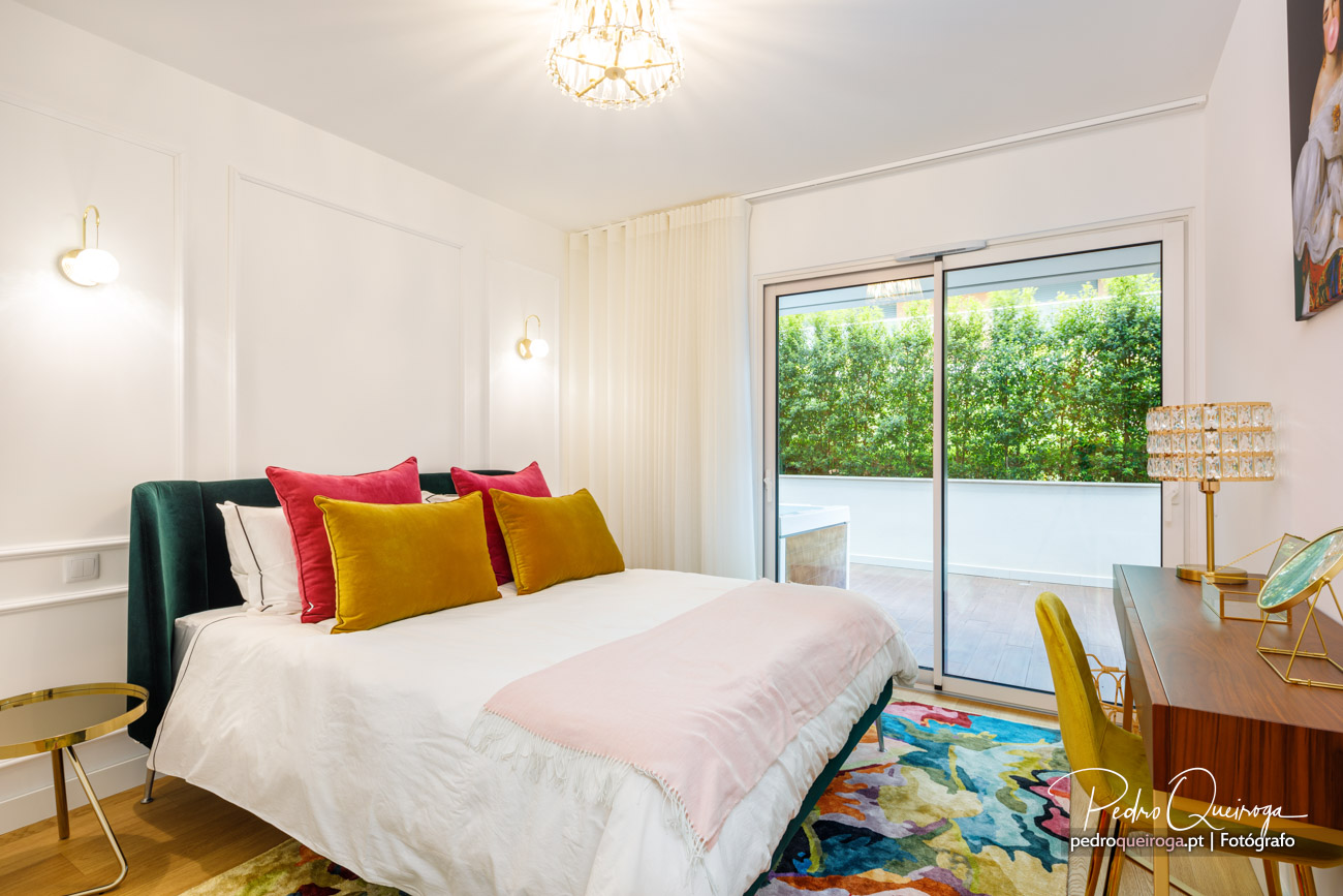 Quarto luminoso com almofadas coloridas, porta de vidro para varanda e detalhes dourados na decoração moderna.