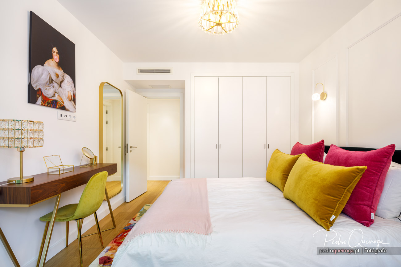 Quarto luminoso com almofadas amarelas e rosa, espelho dourado, secretária em madeira e roupeiro branco embutido