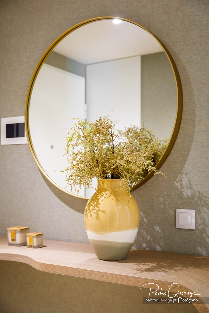 Espelho redondo dourado sobre consola com jarra decorativa amarela e verde com flores secas em parede texturizada bege