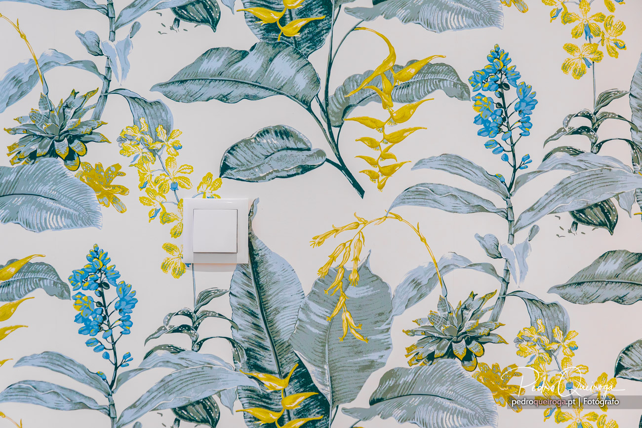 Parede com papel de parede botânico em tons de verde-azulado, amarelo e azul, com interruptor branco ao centro.