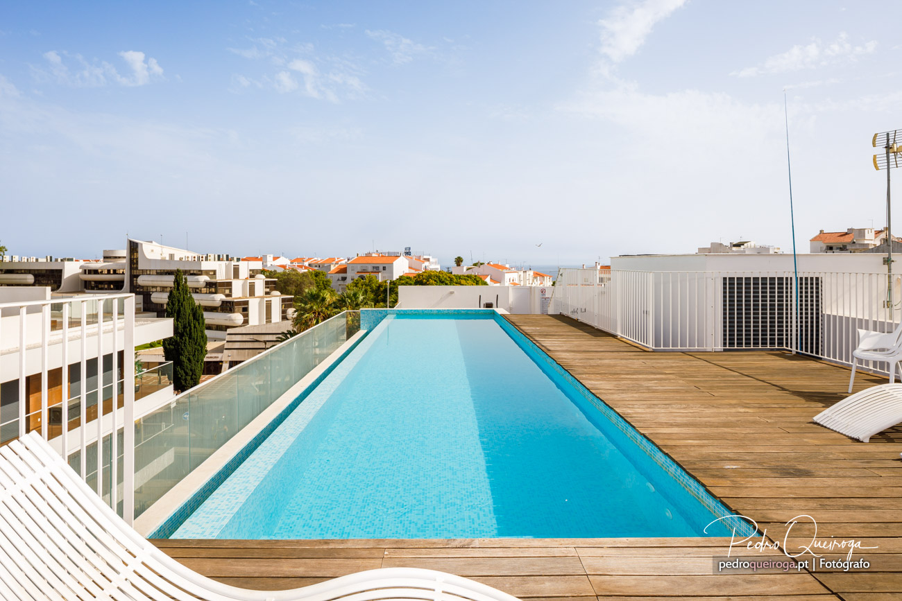 Piscina elegante no rooftop com deck em madeira, vista para telhados laranja e mar ao fundo sob céu azul