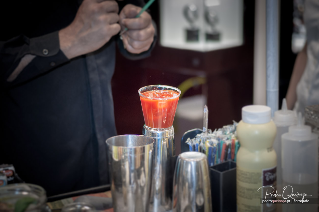 Barman prepara cocktail laranja avermelhado em shaker, rodeado de utensílios profissionais de bar em aço inoxidável