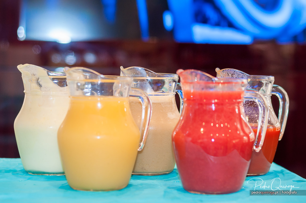 Cinco jarros de sumos naturais coloridos sobre mesa turquesa, com arquitetura de bar desfocada ao fundo.