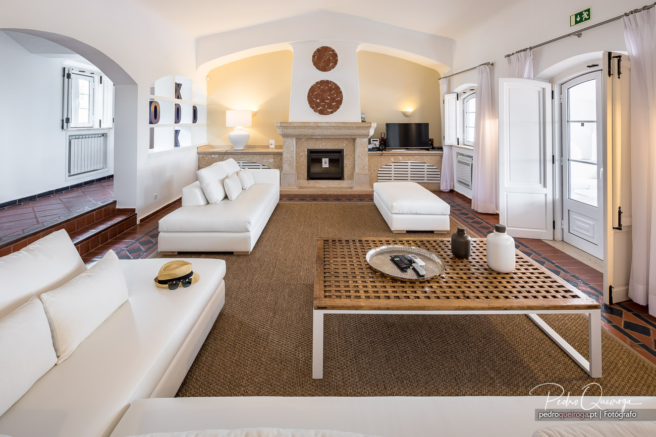 Sala de estar luminosa com sofás brancos, lareira em pedra, decoração circular na parede e portas amplas para o exterior.