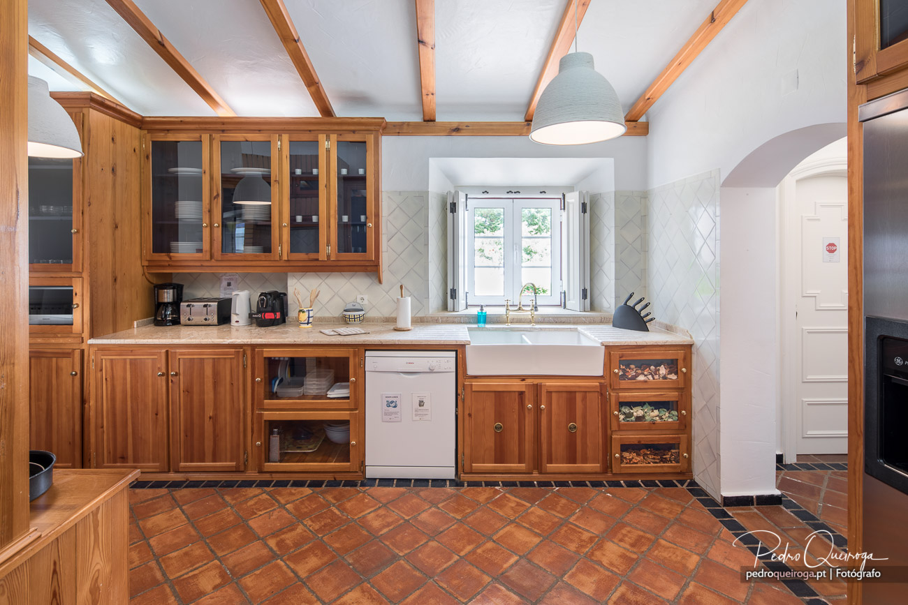 Cozinha ampla com mobiliário em madeira, vigas à vista, bancada em pedra, lava-loiça cerâmica e pavimento terracota