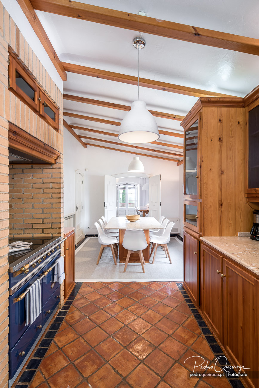 Cozinha rústica com vigas de madeira, pavimento em terracota e sala de jantar ao fundo com cadeiras brancas.