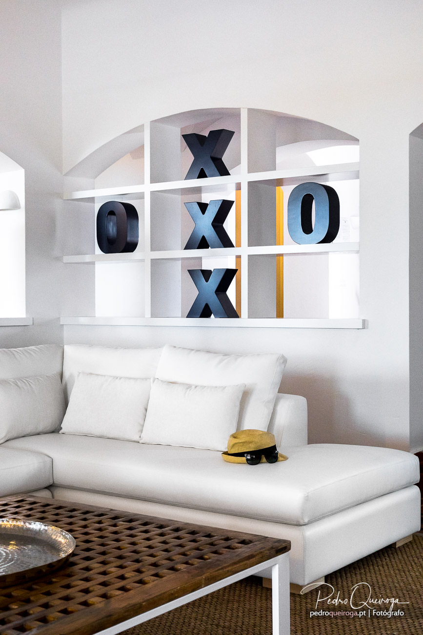 Sala branca minimalista com sofá, estante com letras decorativas em azul e chapéu amarelo sobre almofadas.