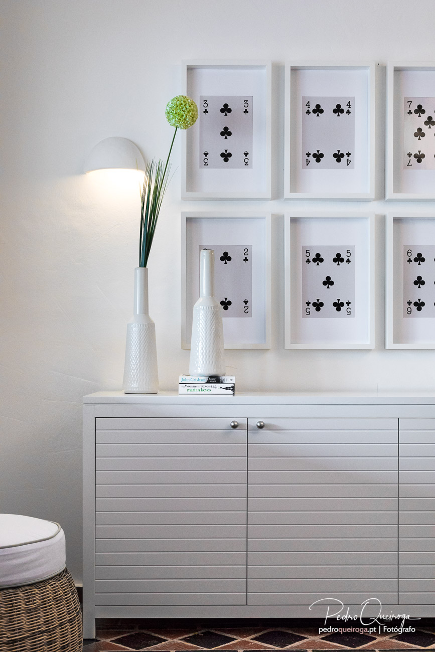 Cómoda cinzenta com candeeiro branco e quadros emoldurados de cartas de paus na parede minimalista