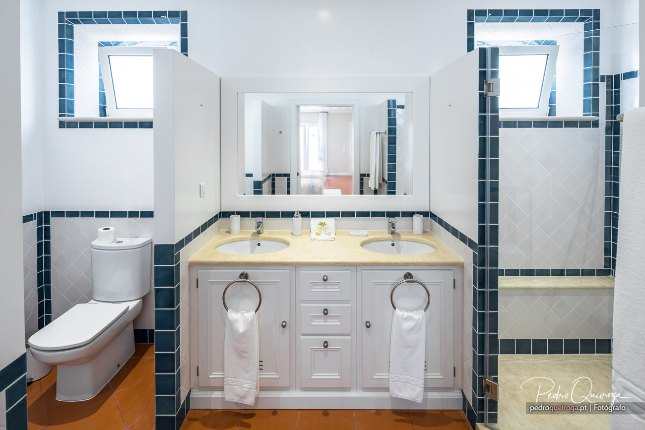 Casa de banho espaçosa com duplo lavatório, bancada amarela, azulejos brancos e detalhes em azul petróleo