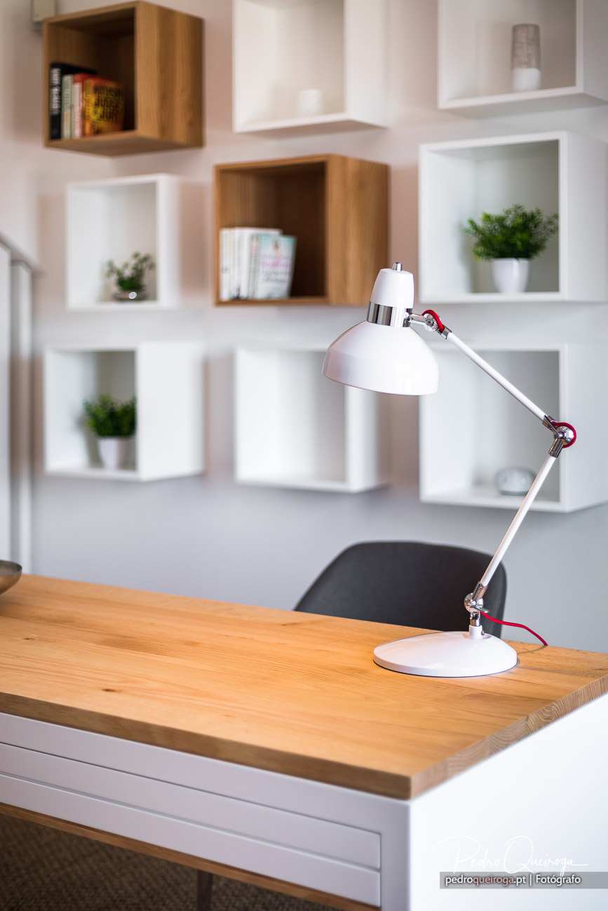 Candeeiro branco articulado sobre secretária de madeira com prateleiras decorativas ao fundo