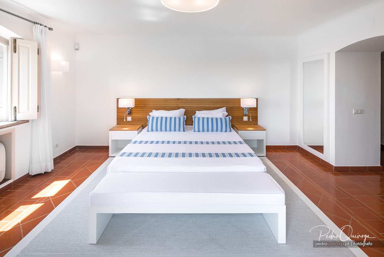 Quarto luminoso com cama de casal, roupa branca e azul às riscas, cabeceira em madeira e chão em terracota.