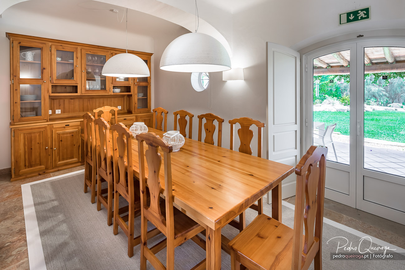 Sala de jantar ampla com mesa e cadeiras em madeira, candeeiros brancos suspensos e vista para o jardim exterior.