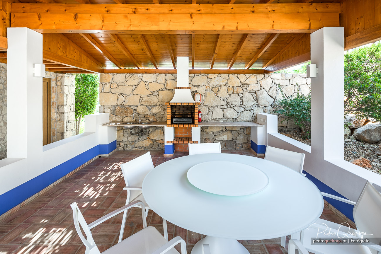 Alpendre coberto com teto em madeira, churrasqueira em tijolo, bancos brancos e azuis, mesa redonda e vegetação envolvente.