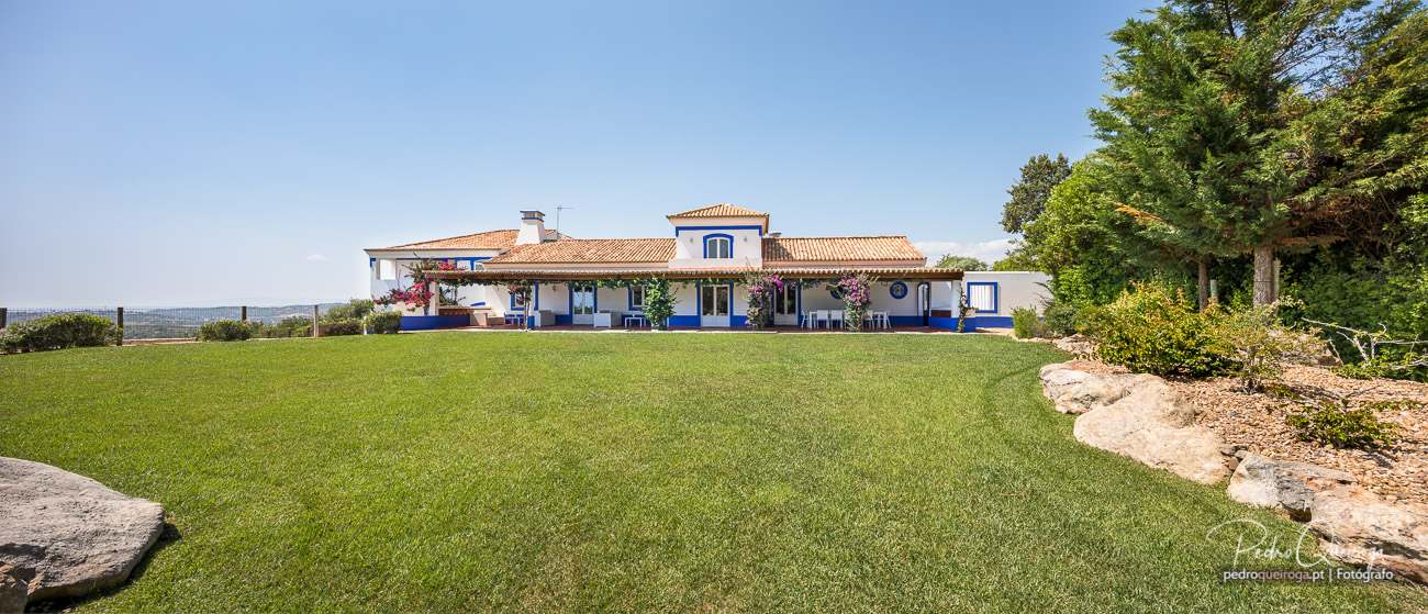 Moradia branca com detalhes azuis, telhado de telha laranja, rodeada por relvado verdejante e árvores sob céu azul claro.