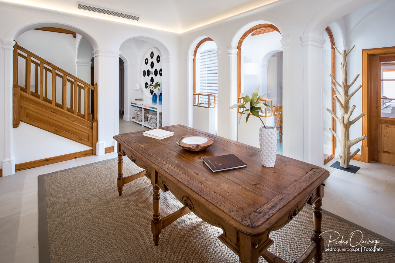 Hall luminoso com arcos brancos, mesa rústica em madeira, cadeiras de fibra e vista para o exterior através de janelas amplas