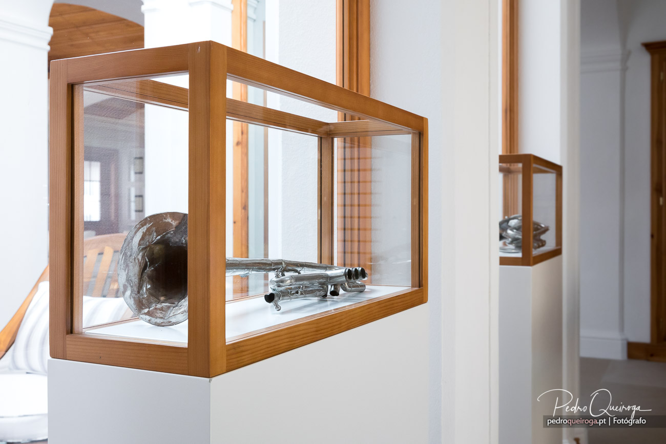 Vitrine em madeira clara com objetos metálicos e escultura escura expostos em galeria moderna e luminosa