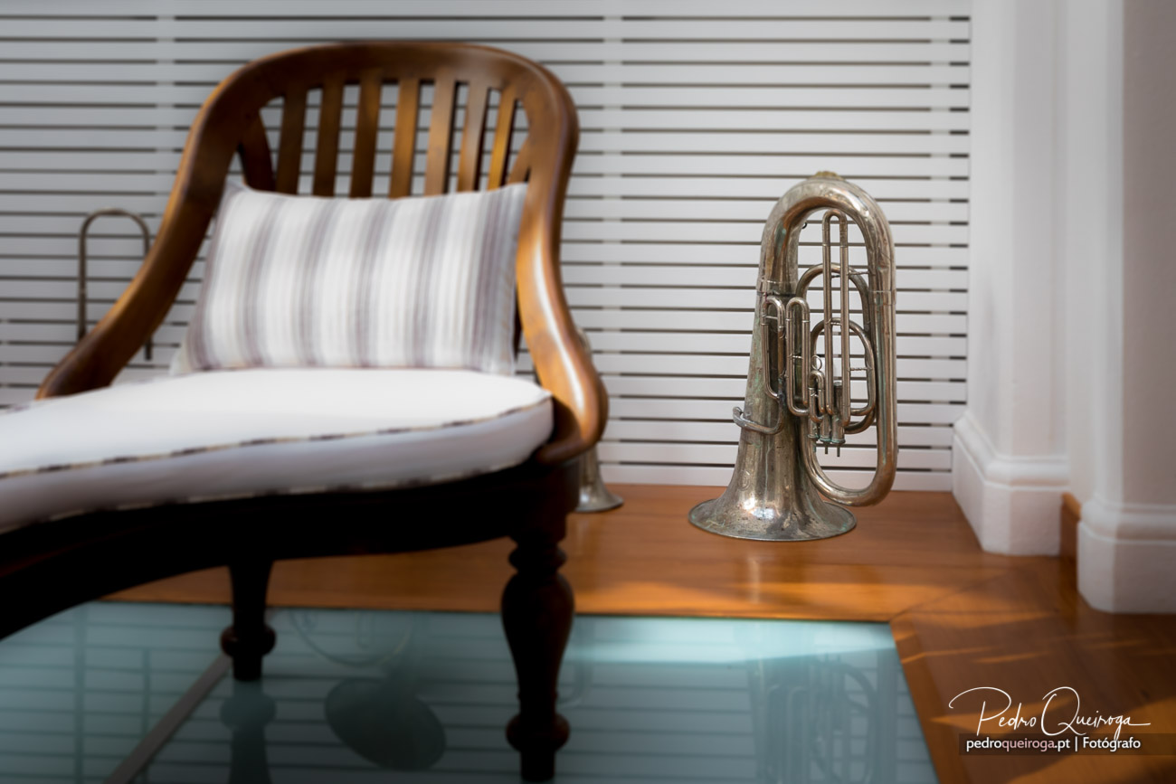 Cama vintage em madeira com almofada às riscas junto a tuba prateada sobre pavimento com tapete azul claro