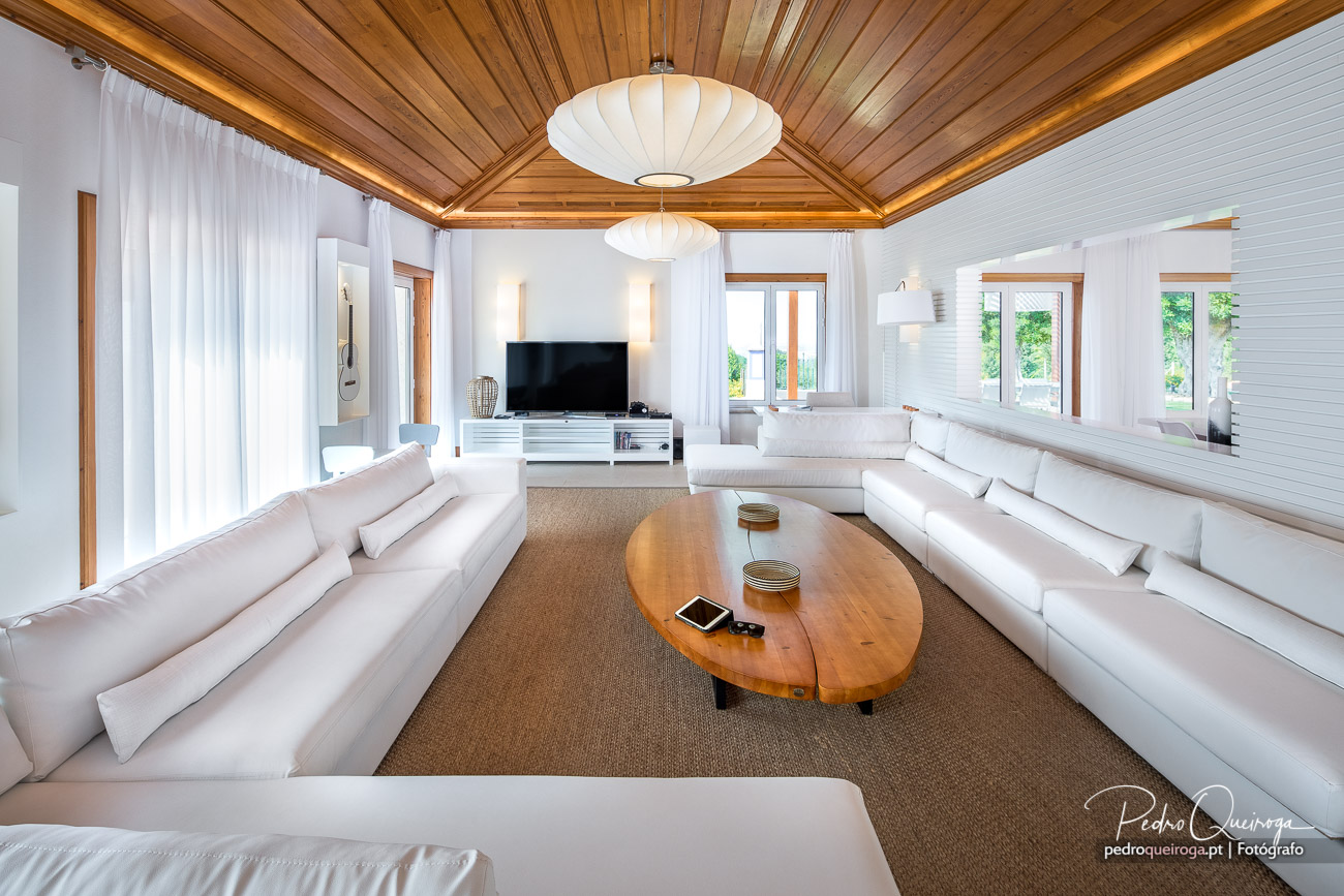 Sala de estar ampla com teto de madeira, sofás brancos, mesa oval central e luz natural abundante das janelas.